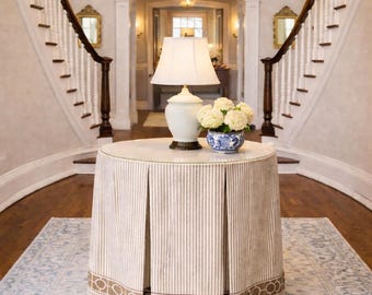 May include: A round accent table with a floor-length, pleated beige and white striped skirt featuring a brown geometric border. On top sits a white ceramic table lamp with a matching shade and a blue and white patterned bowl filled with white flowers.