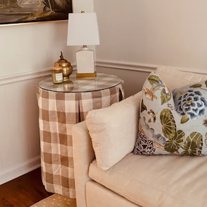 May include: A brown and white checkered side table with a glass top. The table has a gold metal base and is decorated with a gold candle holder and a white lamp with a white shade. The table is next to a white couch with a floral pillow.