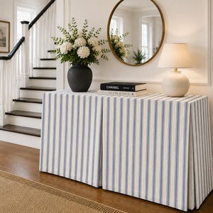 May include: A rectangular console table covered with a floor-length, blue and white striped fabric skirt featuring box pleats. The table surface holds a dark gray vase with white flowers, a white table lamp, and two stacked books with black covers.