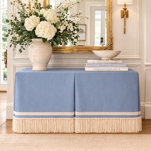 May include: A light blue console table with a cream-colored fringe and trim. The table is topped with a white vase filled with white flowers, books, and a decorative bowl. The text on the books reads "INTERIORS" and "TIMELESS ELEGANCE."