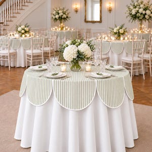 May include: A round table covered with a white floor-length tablecloth and a sage green and white striped scalloped overlay. The table is set with white plates, clear glassware, small candles, and a central bouquet of white hydrangea flowers.
