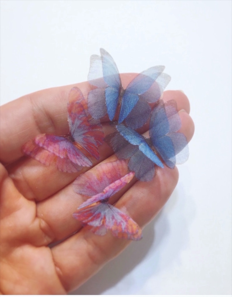 K&ouml;nnte beinhalten: Vier zarte Schmetterlings-Verzierungen in Blaut&ouml;nen und Rosa. Die Schmetterlinge sind aus durchsichtigem Stoff gefertigt und haben eine leicht schimmernde Oberfl&auml;che.