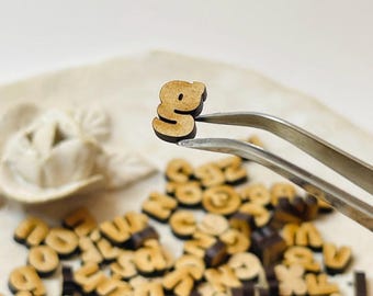 May include: A wooden letter "g" is held by tweezers, with a light brown color and a darker brown outline. A scattering of other wooden letters is in the background, suggesting a craft or educational theme.