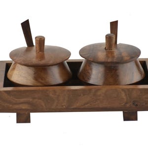 Op de afbeelding: Een houten kruidenset met twee kommen met deksel en kleine lepels, geplaatst in een rechthoekige bak. De kommen en de bak zijn gemaakt van donkerbruin hout, met een zichtbare natuurlijke houtnerf. De set is ontworpen voor het serveren van kruiden of specerijen.
