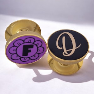 Könnte beinhalten: Zwei goldene Handygriffe mit unterschiedlichen Designs. Ein Griff hat eine lila Blume mit einem schwarzen Buchstaben F in der Mitte. Der andere Griff hat einen schwarzen Hintergrund mit einem goldenen Buchstaben D.