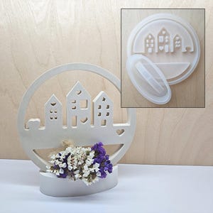 Könnte beinhalten: Ein weißes, kreisförmiges Dekorationselement mit einem Haus-Silhouette-Design, das einen kleinen Strauß Trockenblumen hält. Das Design umfasst ausgeschnittene Fenster und Herzformen. Eine kleinere, transparente Form mit dem gleichen Hausdesign befindet sich in der oberen rechten Ecke.