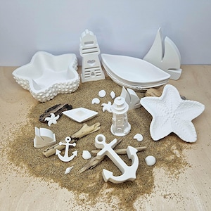 Könnte beinhalten: Eine Sammlung weißer Keramik-Dekorationen zum Thema Nautik, arrangiert auf Sand. Die Artikel umfassen eine sternförmige Schale, ein Leuchtturm, eine Bootsförmige Schale, Anker und Muscheln. Treibholzstücke ergänzen die Präsentation.