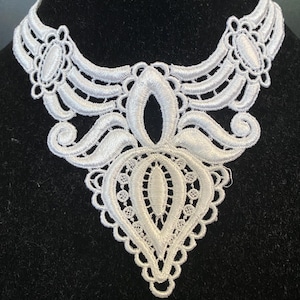 Puede incluir: Un cuello de encaje blanco bordado con un diseño ornamental y simétrico. El cuello presenta un borde festoneado y un elemento central en forma de lágrima. Los intrincados detalles y el calado crean una apariencia delicada y elegante.