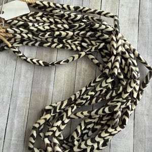 May include: A collection of cream and dark brown beaded strands. The beads have a chevron pattern and are strung together. The strands are tied together with a natural fiber cord. The beads are arranged on a light wood-grain surface.