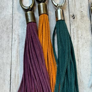 May include: Three suede tassels in purple, orange, and teal, each with a metal clasp. The tassels are hanging against a light wood background. The clasps are in bronze and silver tones.