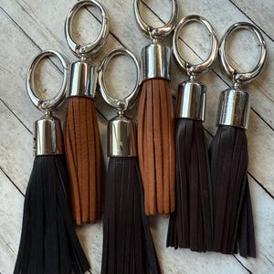 May include: Six keyrings with leather tassels in shades of brown and black. Each keyring has a silver-coloured metal ring and a cylindrical metal cap at the top of the tassel. The tassels are made of fringed leather.