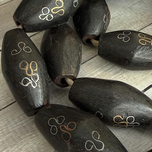 May include: Dark brown wooden beads with a decorative design in silver and gold. The beads are oval-shaped and strung together. The design is a stylized, abstract pattern.