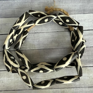 May include: A necklace made of black and white patterned beads. The beads are oval-shaped with a cream-colored base and black geometric designs. The necklace is strung on a natural fiber cord, creating a circular shape.