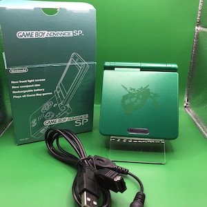 Op de afbeelding: Een groene Game Boy Advance SP console met doos en oplaadkabel. Op de doos staat "GAME BOY ADVANCE SP" en "Nintendo". De console is opengeklapt en toont het scherm. De doos vermeldt ook functies zoals een scherm met voorverlichting en een oplaadbare batterij.