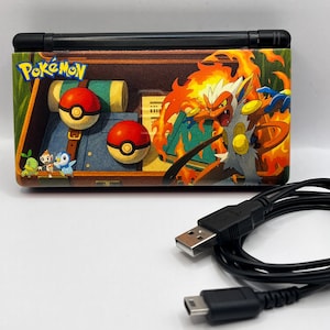 May include: A handheld gaming console with a colorful Pokemon design. The console features a black hinge and a vibrant graphic of Pokemon characters, including a fiery Infernape and Poke Balls. A black USB cable is in the foreground.