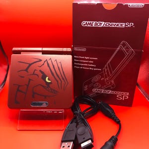 Peut inclure: Une console de jeu portable Game Boy Advance SP rouge avec un graphique de dragon sur le devant. La console est dans sa boîte d'origine, qui présente le logo Game Boy Advance SP et du texte décrivant les caractéristiques de la console. La console est également présentée avec un câble USB et une prise casque.