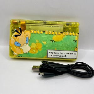May include: A translucent yellow handheld video game console featuring a graphic of the character Psyduck and a row of smaller Psyduck figures. A text box reads, "Psyduck hurt itself in its confusion!" A black USB charging cable is included.