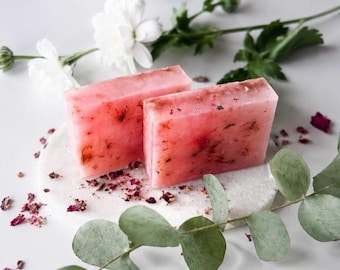 May include: Two rectangular bars of translucent pink soap embedded with dried flower petals. The soap bars are positioned upright on a circular white stone base, surrounded by scattered dried rose petals and green eucalyptus leaves.