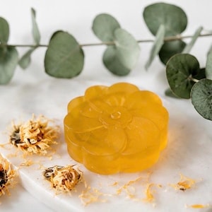 May include: A translucent, golden-yellow, flower-shaped bar of soap with a smooth, moulded petal design. The soap is accompanied by dried yellow flower blossoms and a sprig of green eucalyptus leaves, creating a natural, botanical aesthetic.