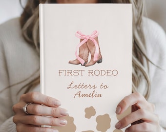 Könnte beinhalten: Ein cremefarbenes Notizbuch mit dem Text "FIRST RODEO Letters to Amelia". Das Cover zeigt eine Aquarellillustration von braunen Cowboy-Stiefeln, die mit einer rosa Schleife gebunden sind. Kuhfellmuster akzentuieren den unteren Bereich.