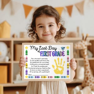 May include: A young child is holding a framed certificate. The certificate says "My Last Day of First Grade" and features a yellow handprint and crayon illustrations. The background includes a wooden shelf and toys.
