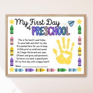 May include: A framed print with the text "My First Day of Preschool." The print features a yellow handprint, colorful crayons, and a poem. The frame is a light brown color.