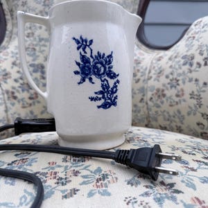 May include: A white ceramic pitcher with a blue floral design. The pitcher has a handle and a spout. A black electrical cord and plug are in the foreground. The pitcher is sitting on a floral patterned chair.