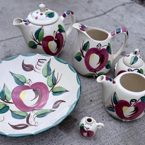 May include: A vintage ceramic tea set featuring a teapot, creamer, sugar bowl, and serving dish. The set is decorated with a hand-painted apple design in shades of red, green, and purple. The handles and rims have a checkered pattern.