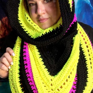 May include: A hooded cowl scarf featuring a crochet pattern with alternating bands of black, neon yellow, and bright pink yarn. The garment is designed with a deep V-shaped drape and a wide, integrated hood that frames the head and neck area.