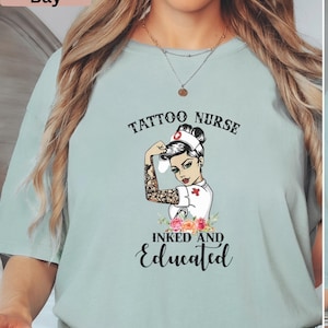 May include: A light blue t-shirt with the words "Tattoo Nurse" above an illustration of a nurse with tattoos and the words "Inked and Educated" below. The nurse illustration is flexing her arm.