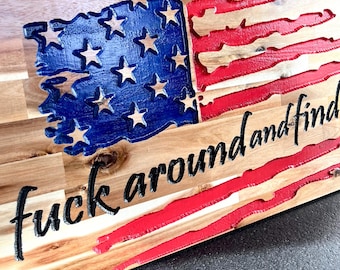 May include: Wooden American flag with a distressed design. The flag features blue stars, red stripes, and the phrase "fuck around and find out" in black lettering. The wood has a natural grain.
