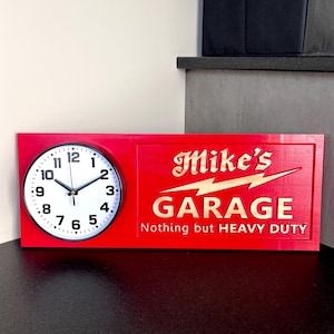May include: A red wooden clock with a white clock face and black hands. The clock is combined with a sign that reads "Mike's GARAGE" in gold lettering, with the text "Nothing but HEAVY DUTY" below.
