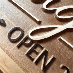 May include: Close-up of a wooden sign with the word "OPEN" carved into the surface. The sign is painted in a light brown color and has a rustic appearance.