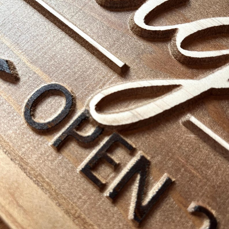 May include: Close-up of a wooden sign with the word "OPEN" carved into the surface. The sign is painted in a light brown color and has a rustic appearance.