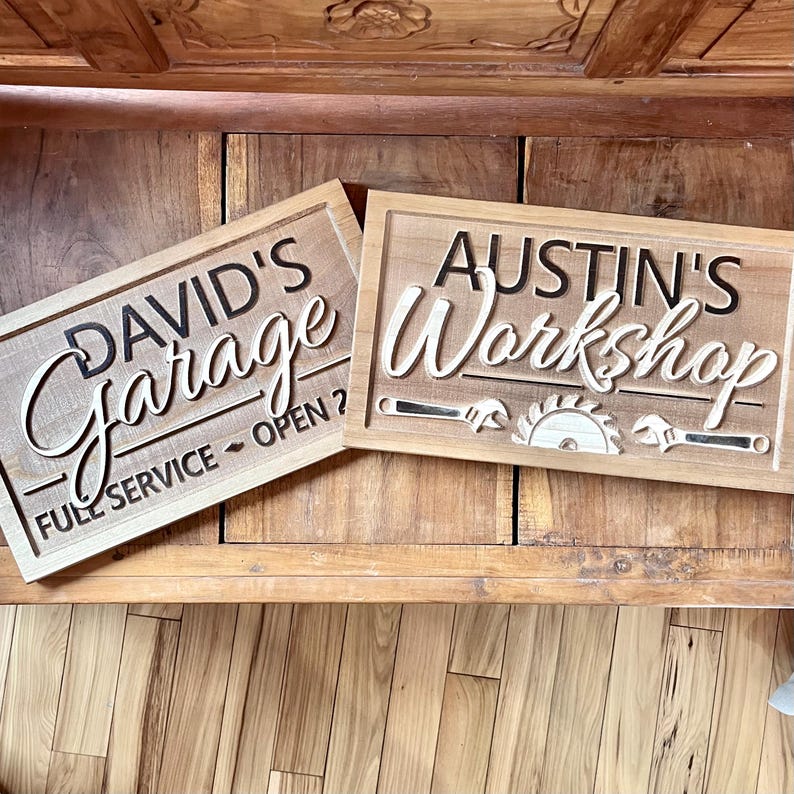 May include: Two wooden signs with carved lettering. The first sign says "David's Garage - Full Service - Open?" The second sign says "Austin's Workshop" with a saw blade and wrench design.