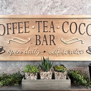 K&ouml;nnte beinhalten: Ein Holzschild mit dem Text "COFFEE - TEA - COCOA - BAR" und den Worten "open daily - self service" in einer kursivschriftlichen Schriftart. Das Schild hat zwei stilisierte, dampfende Kaffeetassen auf beiden Seiten des Textes.