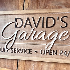 May include: A wooden sign with the text "David's Garage" in black lettering and "Full Service - Open 24/" in black lettering.