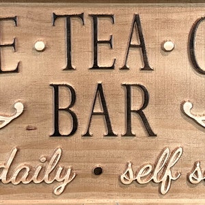 K&ouml;nnte beinhalten: Ein Holzschild mit dem Text "Coffee Tea & Co. Bar" in schwarzen Buchstaben. Das Schild tr&auml;gt auch den Text "Open daily. Self service" in einer kursivschriftlichen Schrift.