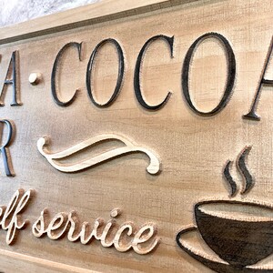 K&ouml;nnte beinhalten: Ein Holzschild mit dem Text "Tea - Cocoa Bar - self service" in schwarzen Buchstaben. Das Schild zeigt auch ein stilisiertes Design einer Kaffeetasse mit aufsteigendem Dampf.