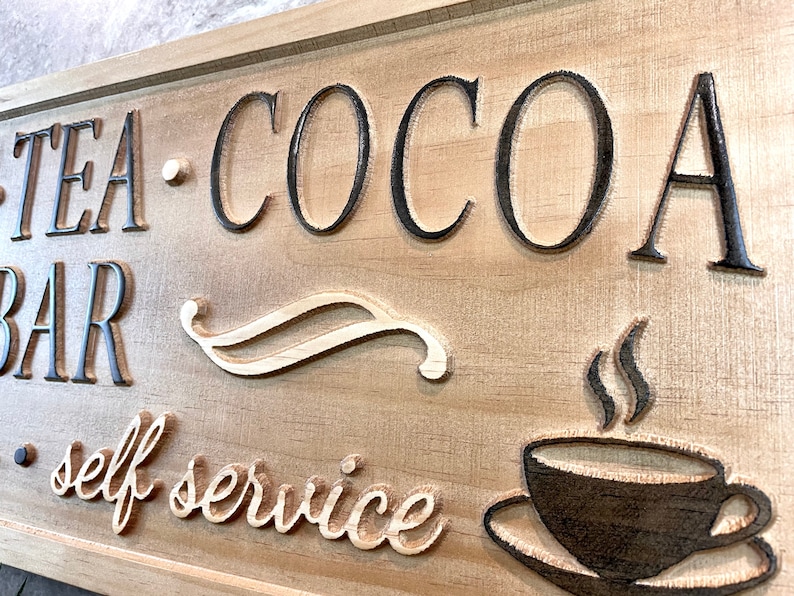 K&ouml;nnte beinhalten: Ein Holzschild mit dem Text "Tea - Cocoa Bar - self service" in schwarzen Buchstaben. Das Schild zeigt auch ein stilisiertes Design einer Kaffeetasse mit aufsteigendem Dampf.