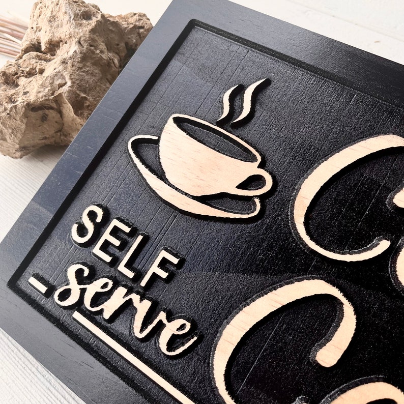 May include: Black wooden sign with white lettering that reads "Self-Serve Coffee". The sign features a stylised image of a coffee cup with steam rising from it.