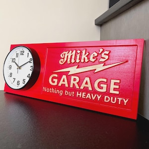 May include: A rectangular red wall sign featuring a circular analog clock on the left side with a white face and black numerals. The sign displays the text Mike's GARAGE Nothing but HEAVY DUTY in raised light-colored lettering with a lightning bolt.