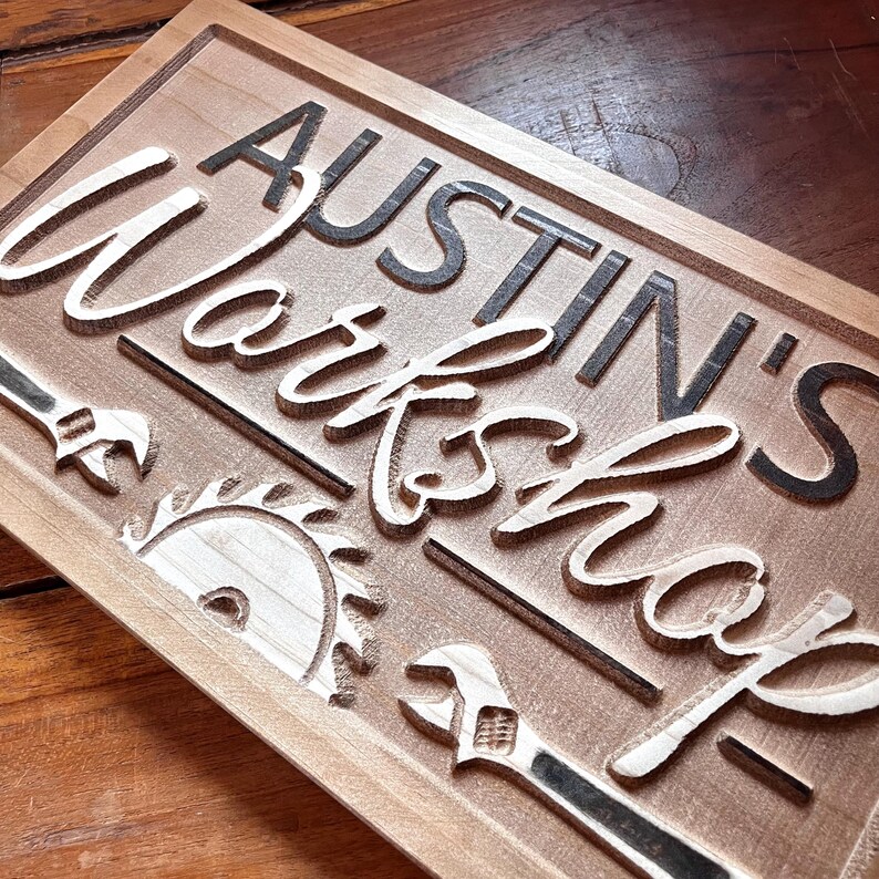 May include: A wooden sign with the text "Austin's Workshop" in black lettering. The sign is decorated with a saw blade and wrench.