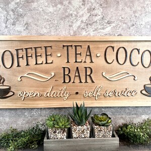 K&ouml;nnte beinhalten: Ein Holzschild mit dem Text "Coffee Tea Cocoa Bar" und den Worten "open daily - self service". Das Schild ist braun und hat einen rustikalen Look. Auf dem Schild sind zwei Kaffeetassen mit Dampf zu sehen.