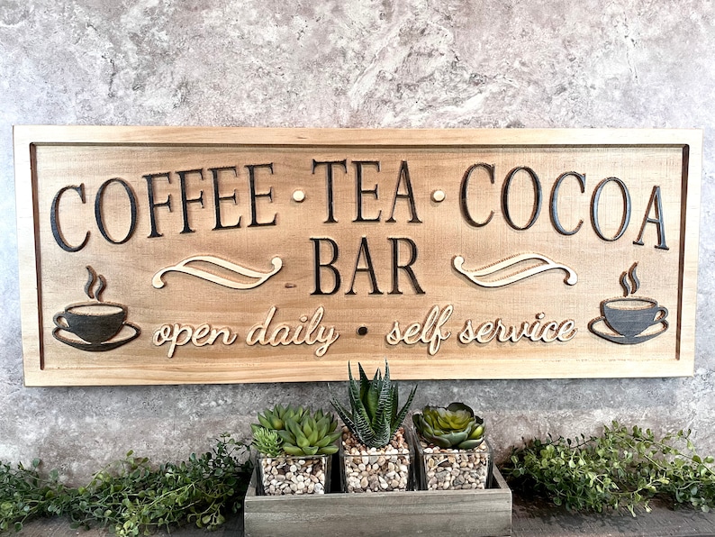 K&ouml;nnte beinhalten: Ein Holzschild mit dem Text "Coffee Tea Cocoa Bar" und den Worten "open daily - self service". Das Schild ist braun und hat einen rustikalen Look. Auf dem Schild sind zwei Kaffeetassen mit Dampf zu sehen.
