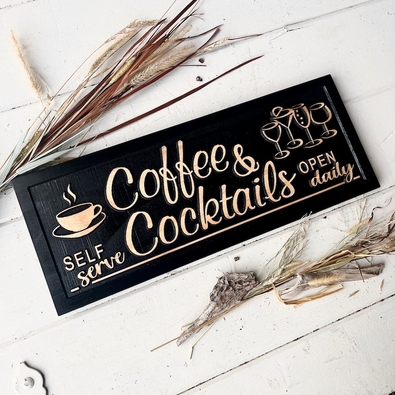 May include: Black wooden sign with gold lettering that reads "Coffee & Cocktails Open Daily" and "Self Serve". The sign also features a gold silhouette of a coffee cup and a group of cocktail glasses.