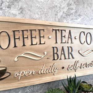 K&ouml;nnte beinhalten: Ein Holzschild mit dem Text "COFFEE - TEA - COCOA BAR" und "open daily - self service". Das Schild ist braun mit schwarzer Beschriftung und einem wei&szlig;en Design.