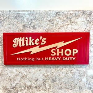 May include: Red wooden sign with the text "Mike's Shop" and the words "Nothing but HEAVY DUTY" in gold lettering. A lightning bolt graphic is above the word "Shop".