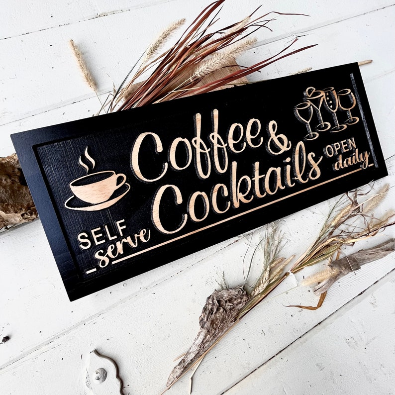 May include: Black wooden sign with the text "Coffee & Cocktails" in gold lettering. The sign also says "Self Serve" and "Open Daily". There is a small coffee cup icon and a group of four cocktail glass icons.
