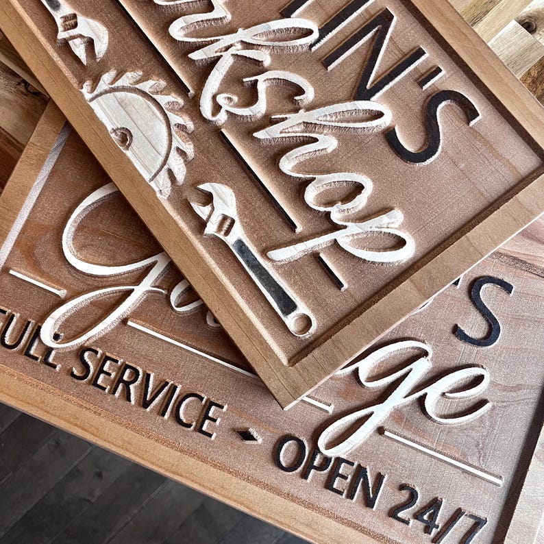 May include: Two wooden signs with white lettering. The top sign says "IN'S Workshop" with a saw blade and wrench design. The bottom sign says "Garage Open 24/7" with a white "G" and "Open 24/7" in black lettering.
